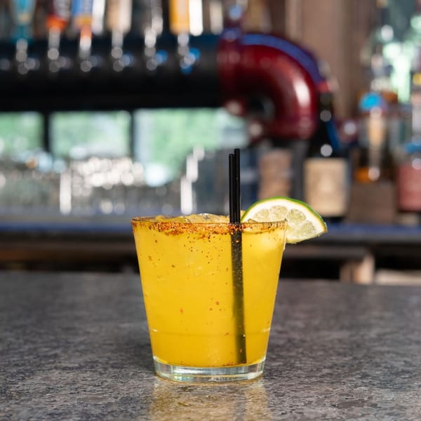 Yellow cocktail with a chili-lime rim, lime wedge, and straws on a bar counter with taps in the background.