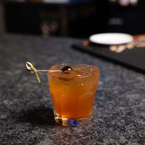 An amber cocktail with ice and a cherry garnish on a dark bar counter.