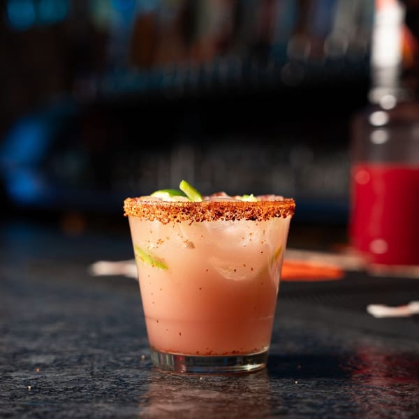 Pink cocktail with a chili-salt rim and lime wedges on a dark bar counter.