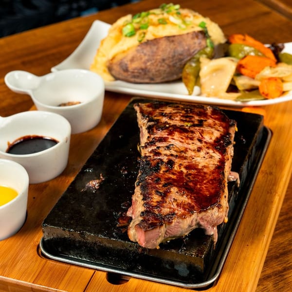 Grilled steak on a hot stone with a baked potato, mixed vegetables, and dipping sauces.
